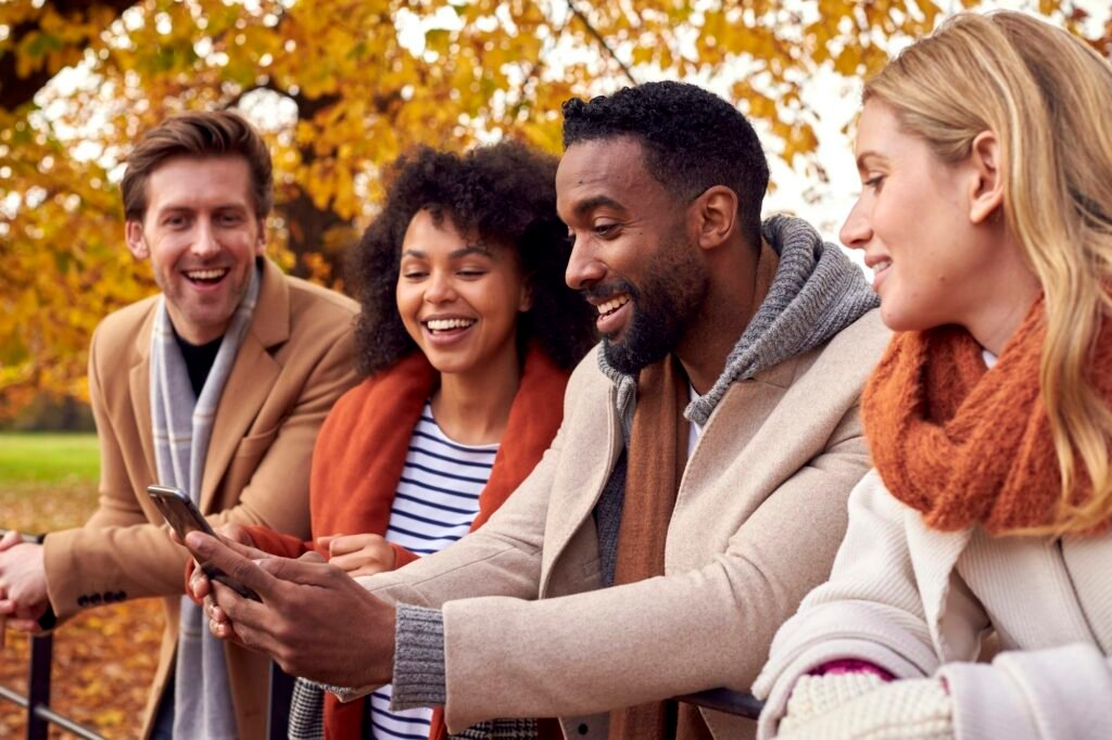 Group Of Friends Outdoors Wearing Coats And Scarves Looking At Photos On Phone In Autumn Park